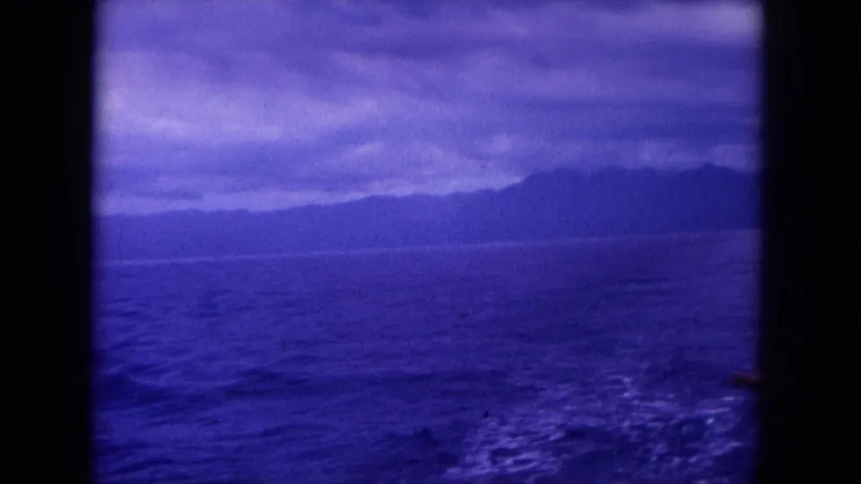 Likely Hawaii coastline from a moving boat in 1964 home-movie footage, with wake patterns, open water, and dark landforms on the horizon.