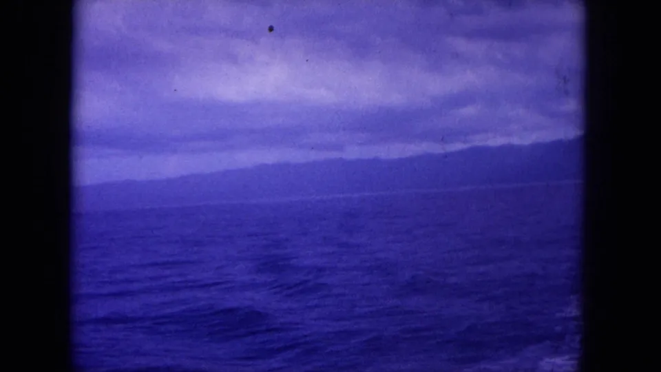 1964 archival home-movie frame likely near Hawaii, showing open sea, a low distant shoreline, cloudy sky, and visible 8mm frame edges.
