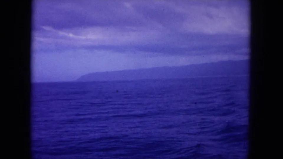 End frame from likely Hawaii 1964 8mm boat footage, with rippled ocean, hazy coastline, cloud cover, and strong blue-purple film color.