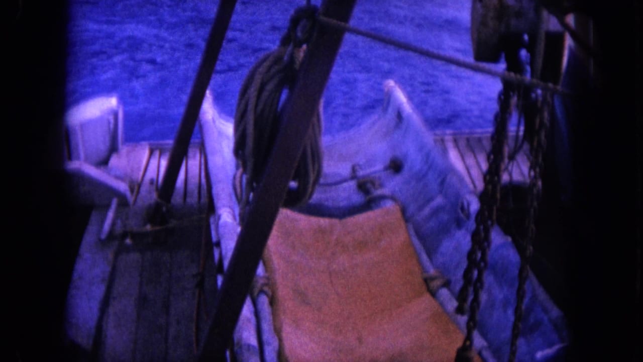 Boat deck with a wooden sling chair, ropes, chain, railings, and rigging above blue water.
