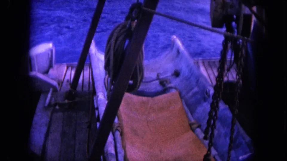 1964 Hawaii 8mm frame of a fishing boat deck with a weathered wooden sling chair, rope coils, chain, railings, and rigging.