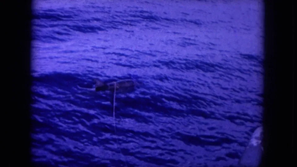 Blue-tinted 1964 Hawaii home-movie frame showing rippled ocean water and a fishing line leading to a fish near the surface.