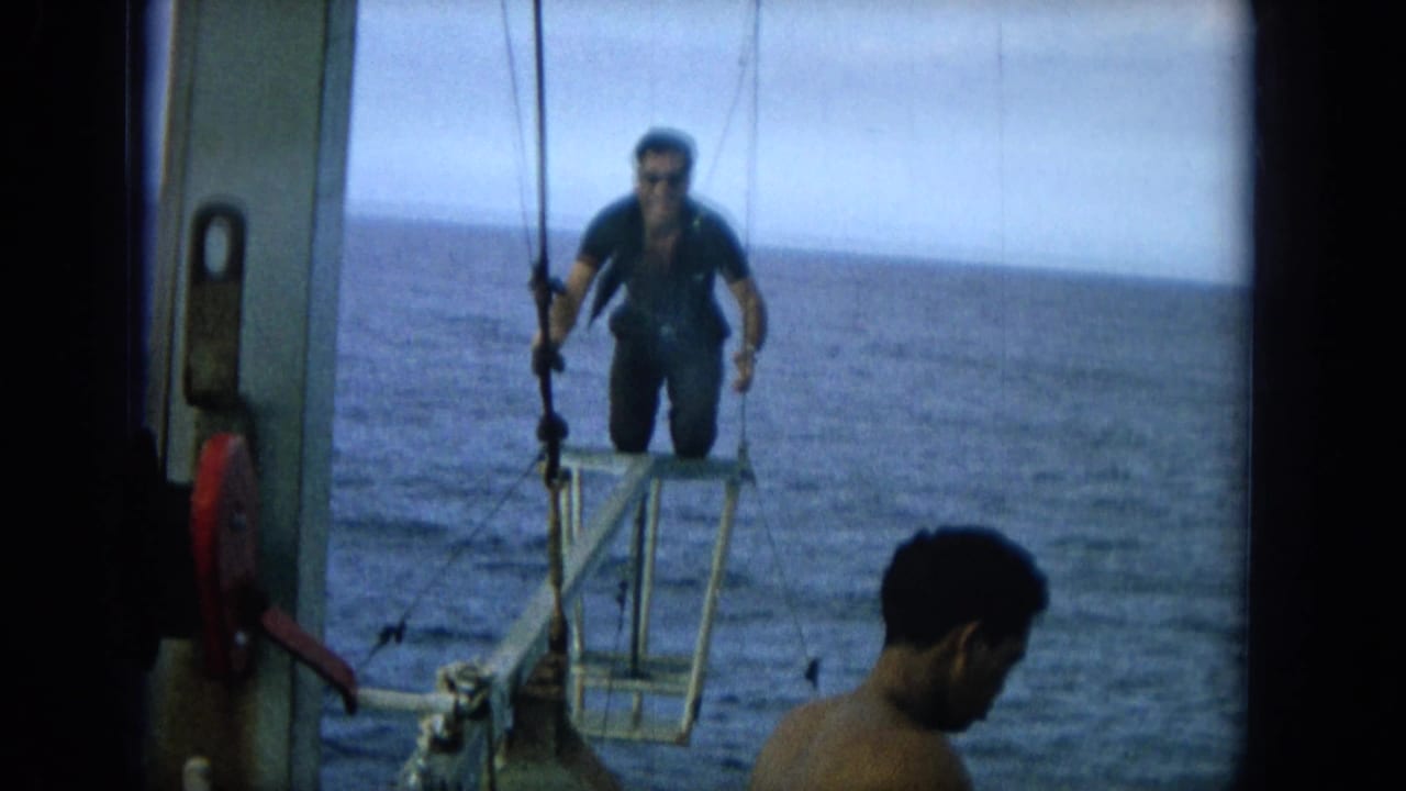 1964 Hawaii home-movie frame with a man in a dark wetsuit on a small bow platform while another man watches from the boat.