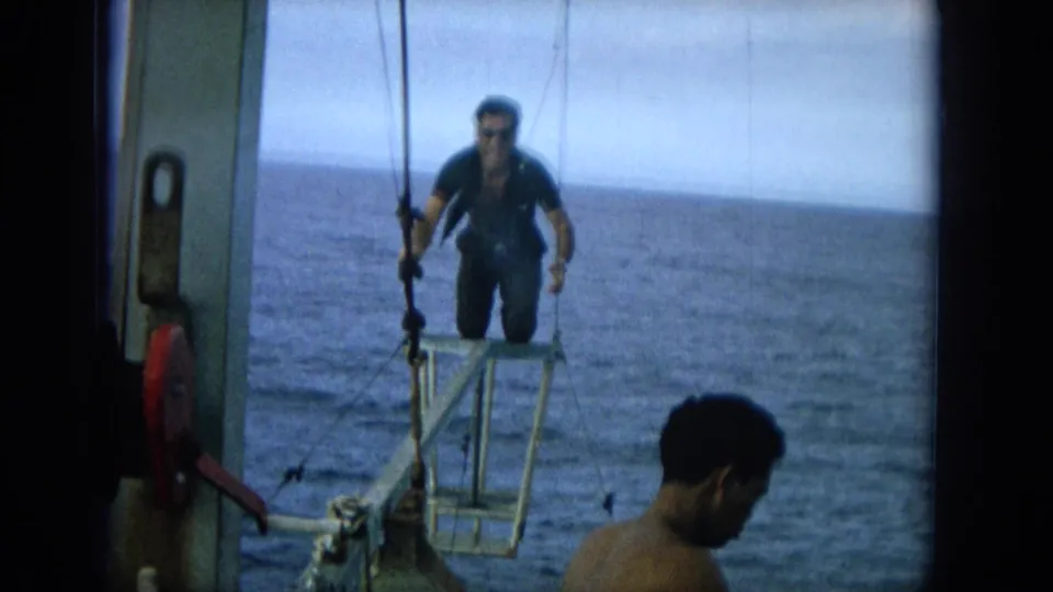 1964 Hawaii home-movie frame with a man in a dark wetsuit on a small bow platform while another man watches from the boat.