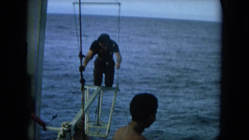 1964 Hawaii 8mm frame showing a wetsuited man bending on the bow platform with open ocean behind him and a passenger in the foreground.