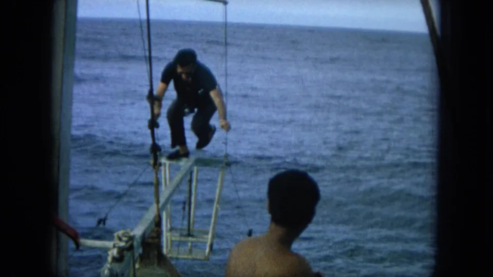 1964 Hawaii home-movie frame with a wetsuited man crouching on the bow platform over the ocean as a passenger looks on.