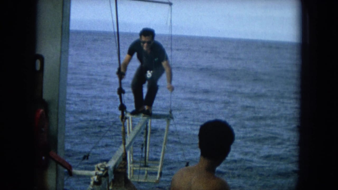 Man in dark clothing balances on a narrow boat platform over the ocean while holding ropes.