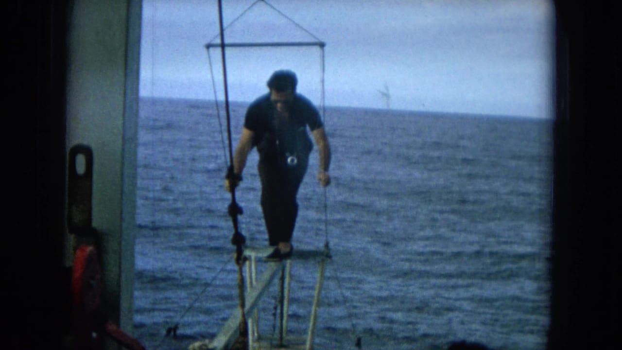 A man in dark clothing stands on a small metal platform extending from a boat over the ocean in supported 1964 Hawaii home-movie footage.