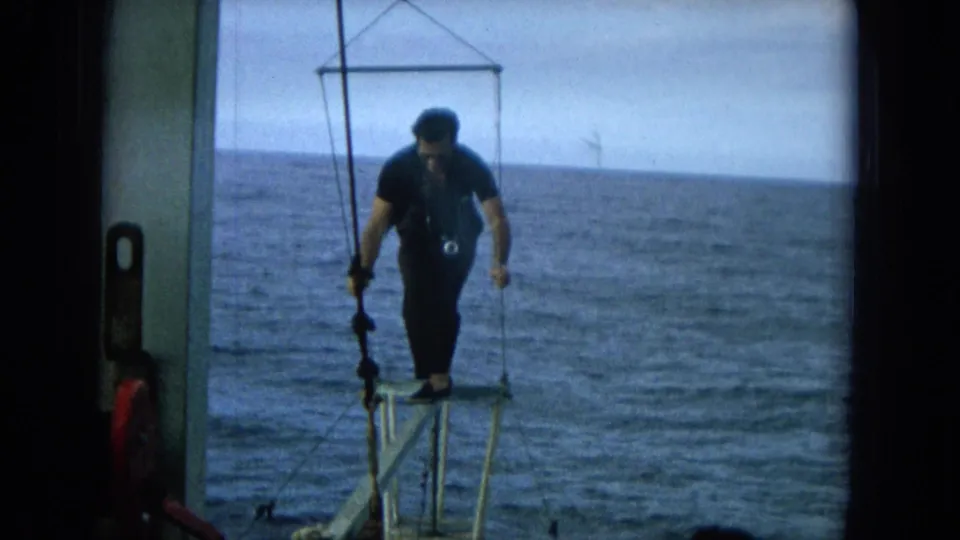 A man in dark clothing stands on a small metal platform extending from a boat over the ocean in supported 1964 Hawaii home-movie footage.