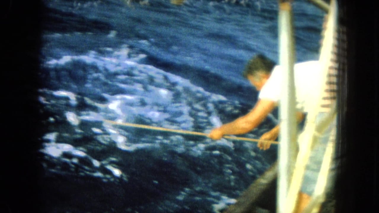Man in a white shirt leans over a boat rail with a rope or line.