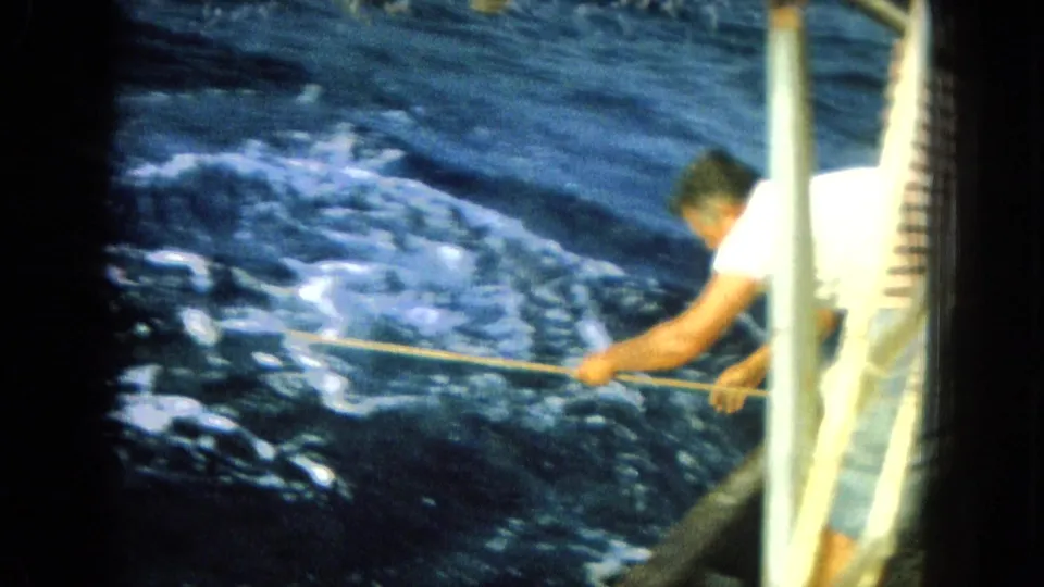 A man in a white T-shirt leans from the boat rail toward the water while handling a rope or line in Hawaii, 1964.