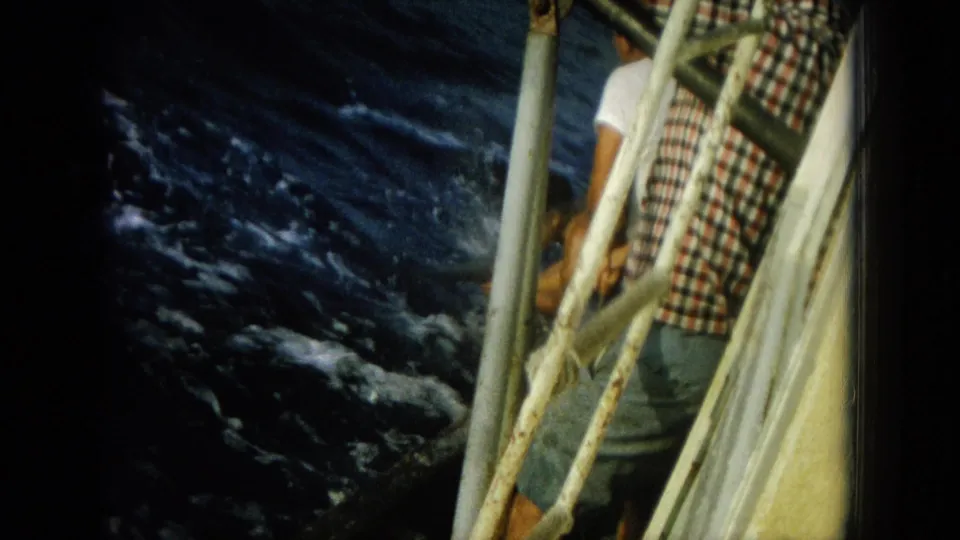 Two visible people stand by the rail of a small boat as the dark ocean moves below in 1964 Hawaii home-movie footage.