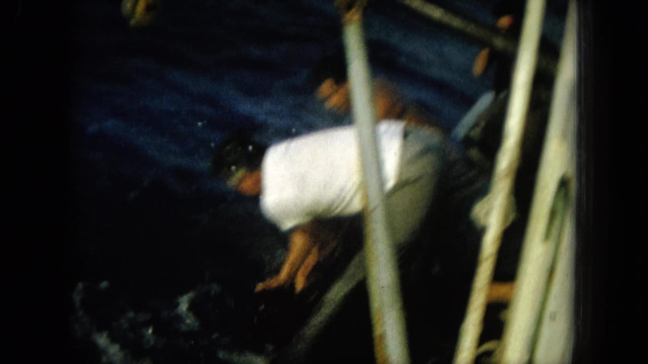In a 1964 Hawaii 8mm home-movie frame, adult men in light clothing lean over a small boat as dark ocean water churns below.