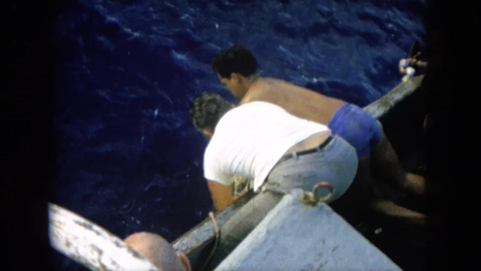 Two adult men in casual clothing lean over the edge of a small boat in the 1964 Hawaii footage, looking down toward the water where the fishing action continues.