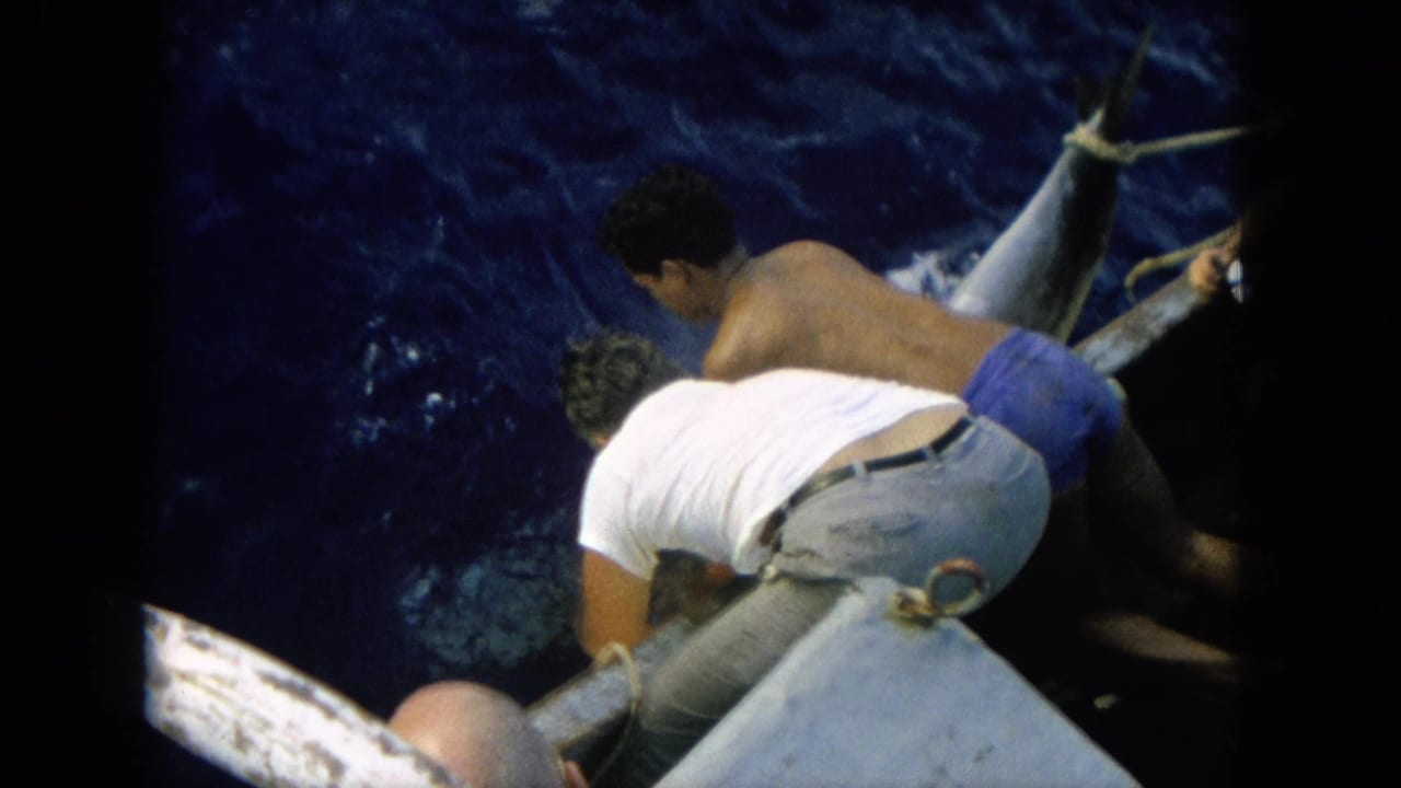 1964 Hawaii 8mm frame showing two adult men working over a boat edge to control a large marlin in deep blue water.