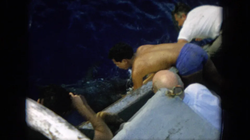 1964 Hawaii 8mm frame with a shirtless man in blue shorts stretched over the boat edge during the marlin catch.