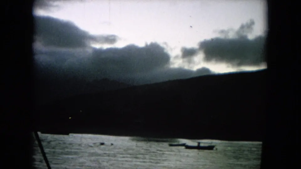 1964 Hawaii home-movie frame showing a cloudy coastal shoreline, calm water, and small boats in the distance.