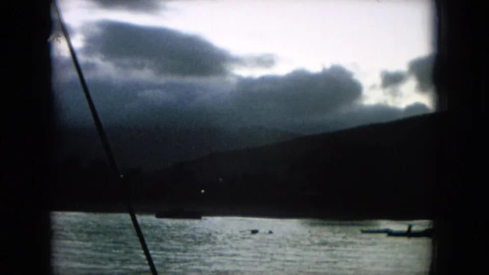 Handheld 8mm view of a Hawaii shoreline under clouds, with water, small boats, and boat hardware crossing the frame.