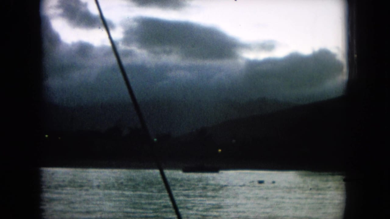 1964 8mm frame identified as Oahi, Hawaii, showing water, distant hills, heavy clouds, and boat rigging in low light.