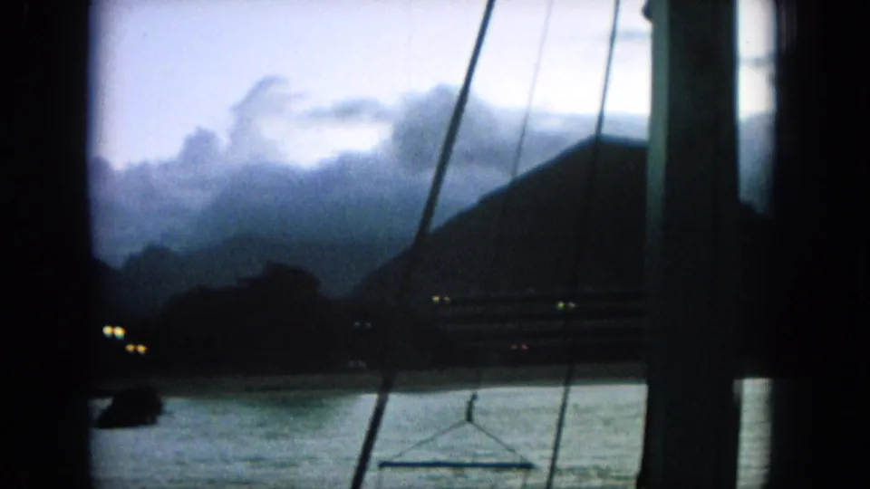 1964 8mm frame identified as Oahi, Hawaii, showing dusk water and distant hills through boat rigging under a cloudy sky.