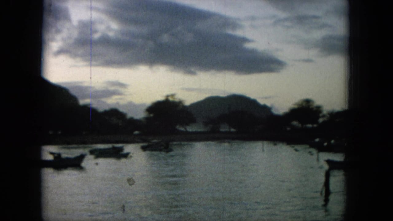1964 8mm Hawaii home-movie frame showing small boats on a dim bay below mountain silhouettes and a cloudy dusk sky.