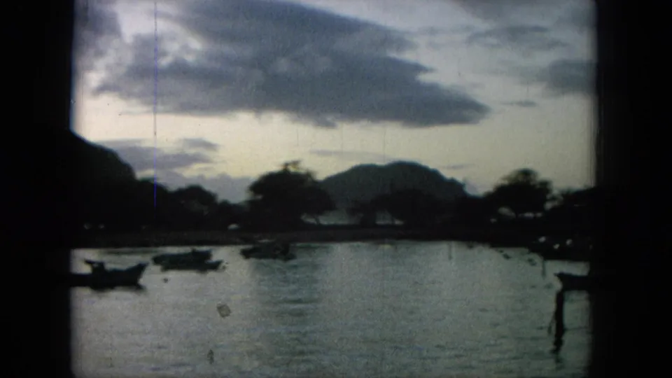 1964 8mm Hawaii home-movie frame showing small boats on a dim bay below mountain silhouettes and a cloudy dusk sky.