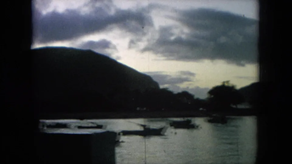 1964 8mm Hawaii frame with anchored boats in a bay and a large dark mountain mass under clouded evening light.