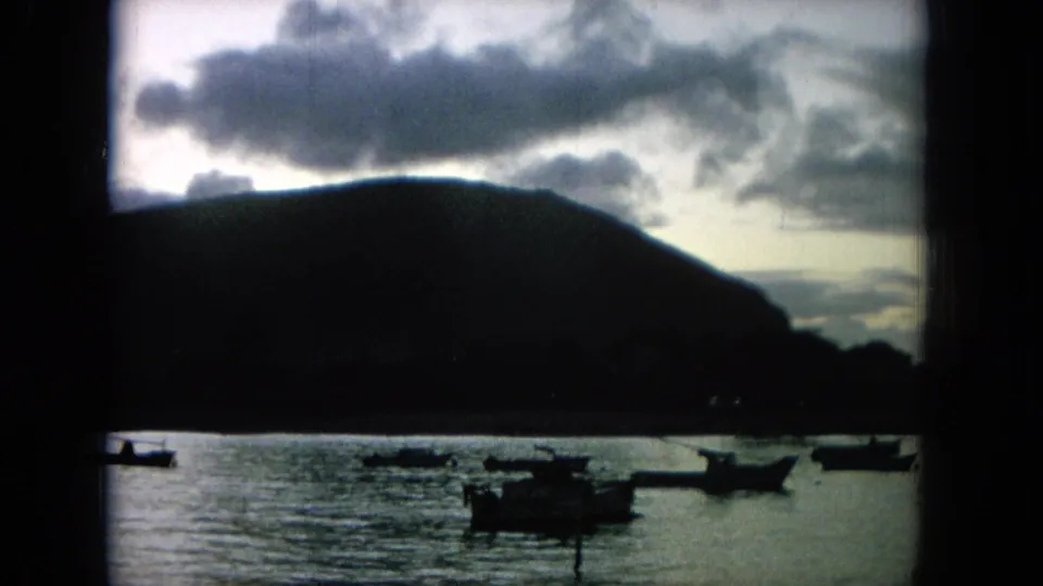 1964 Hawaii home-movie frame showing several small boats across rippled bay water, with a dark mountain ridge and bright cloud cover above.