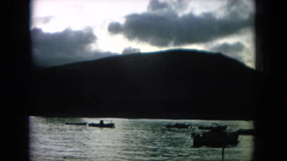 1964 8mm Hawaii frame with boat silhouettes on a bay, a dark mountain slope, and heavy clouds near the horizon.