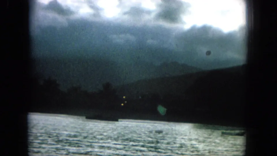 A late frame from the 1964 Hawaii clip shows rippling water, distant boats, dark hillside silhouettes, and layered clouds.