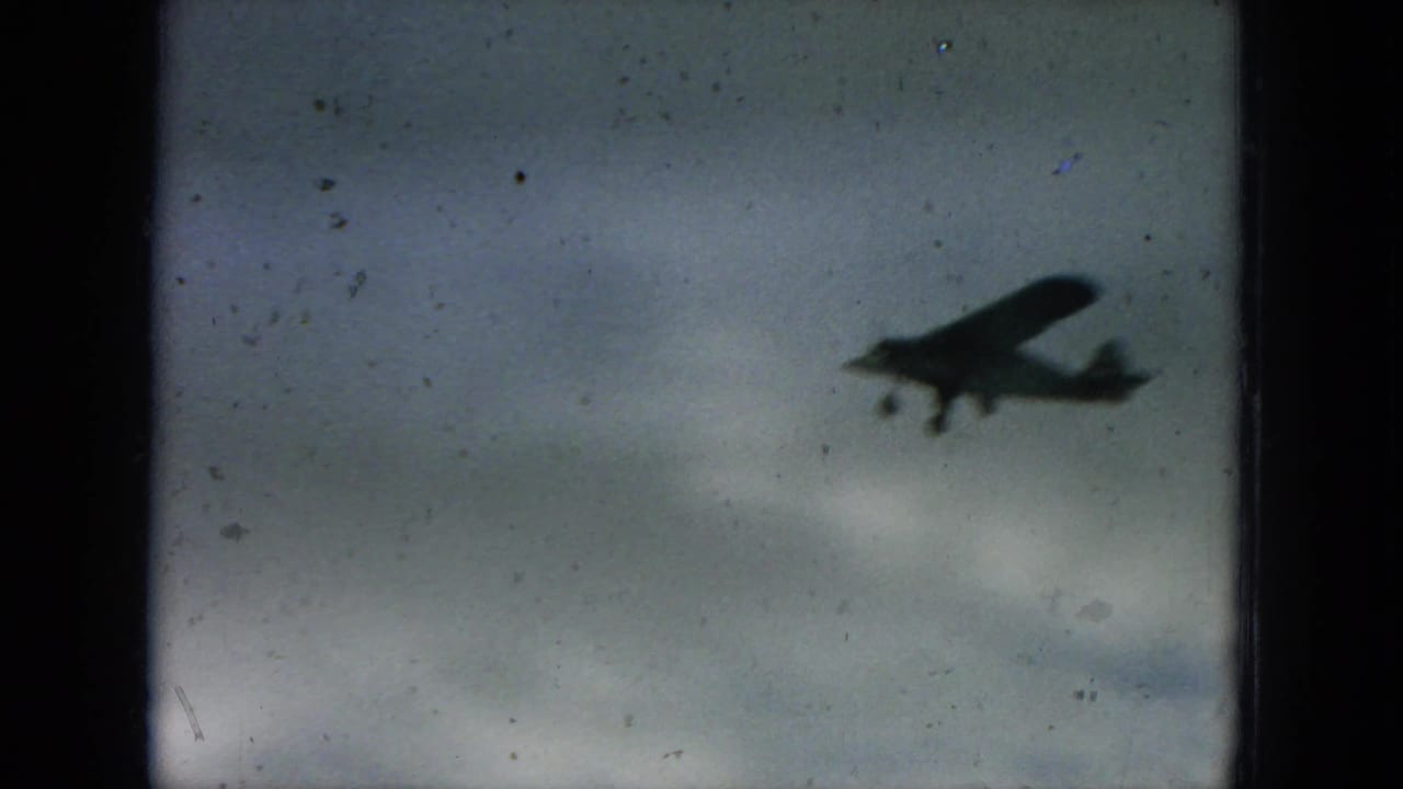Small propeller plane appears in silhouette against a gray sky in this 1964 Hawaii 8mm home-movie frame.