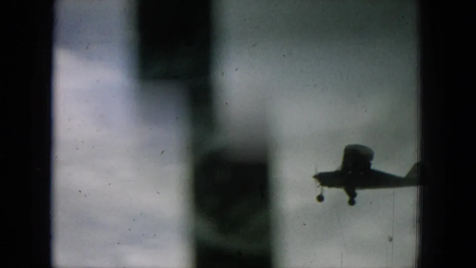 A small plane passes through a soft, dark-framed 1964 Hawaii home-movie view with blurred foreground shapes.