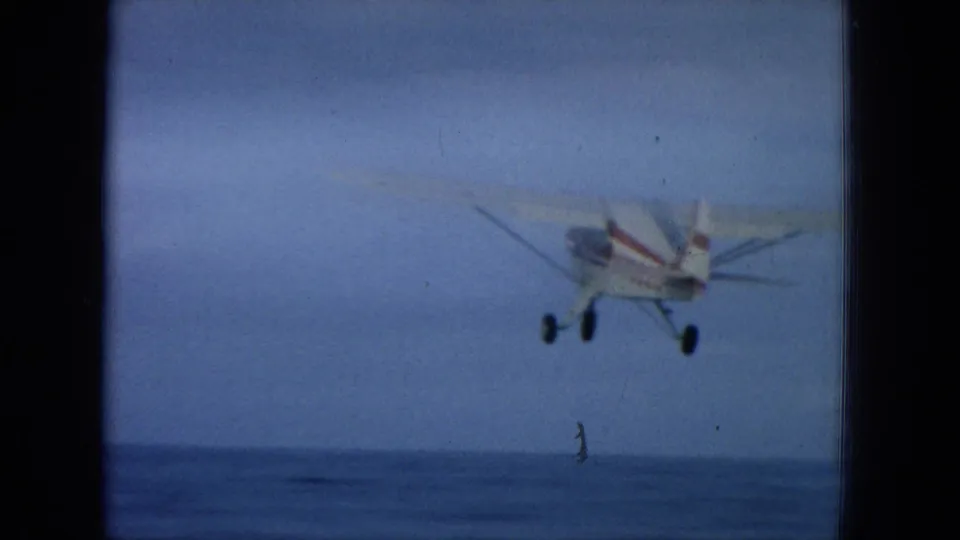 1964 Oahi, Hawaii home-movie frame of a small red-and-white aircraft flying low over the ocean horizon, seen from a moving vessel.