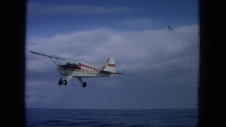 1964 Oahi, Hawaii frame showing a small red-and-white aircraft flying past over the ocean, with visible registration markings on the fuselage.
