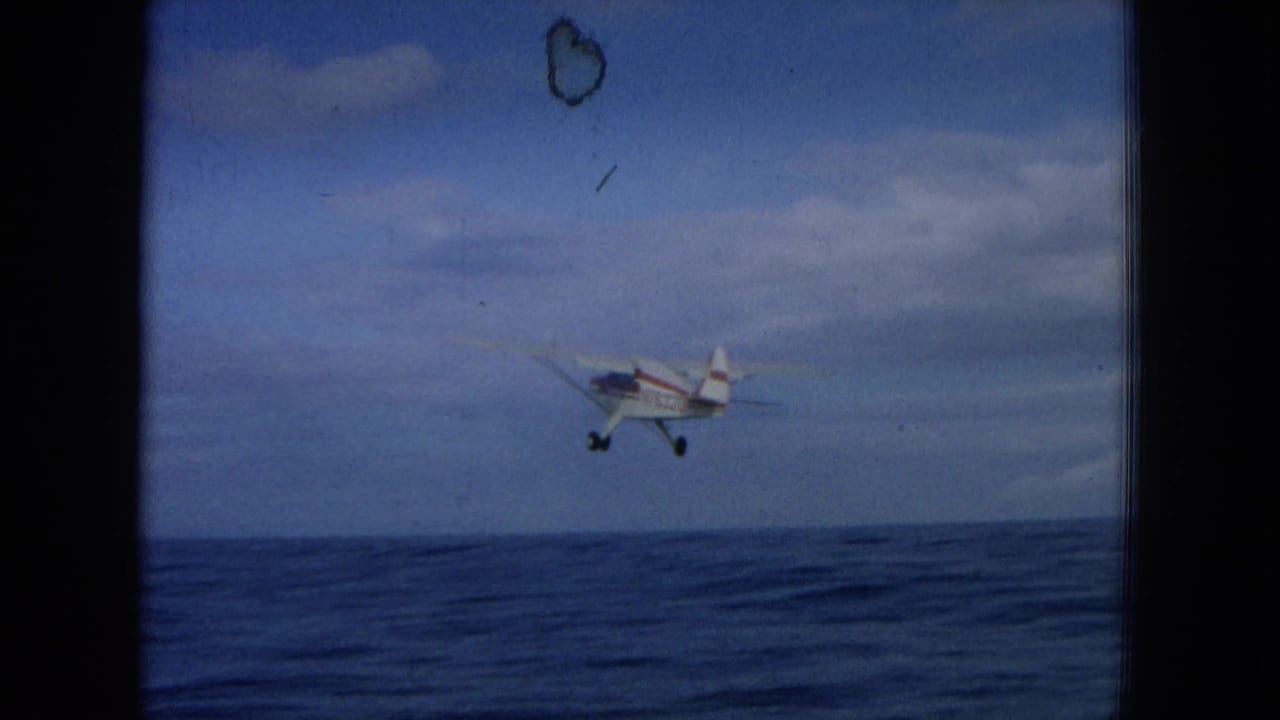 Small white and red plane flying low above the ocean under a cloudy blue sky.
