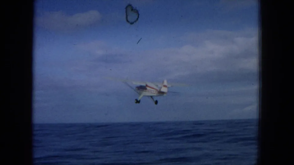 1964 Hawaii 8mm frame showing a small white and red single-engine plane approaching low over open ocean.