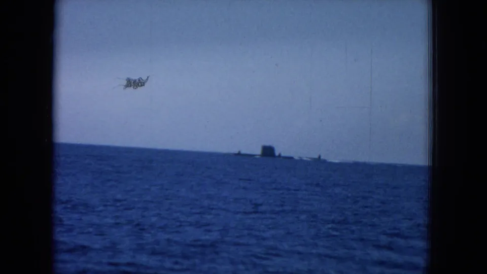 1964 home-movie frame showing blue ocean, a distant ship or submarine-like form, and noticeable film dust in the sky area.