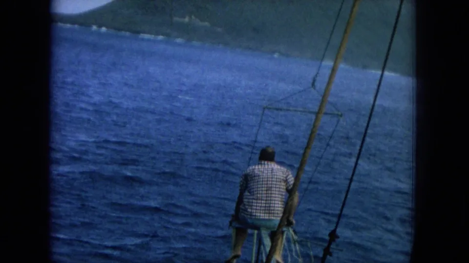 1964 Hawaii 8mm frame with a man in a checkered shirt seated near boat rigging, facing blue water and a green shoreline.