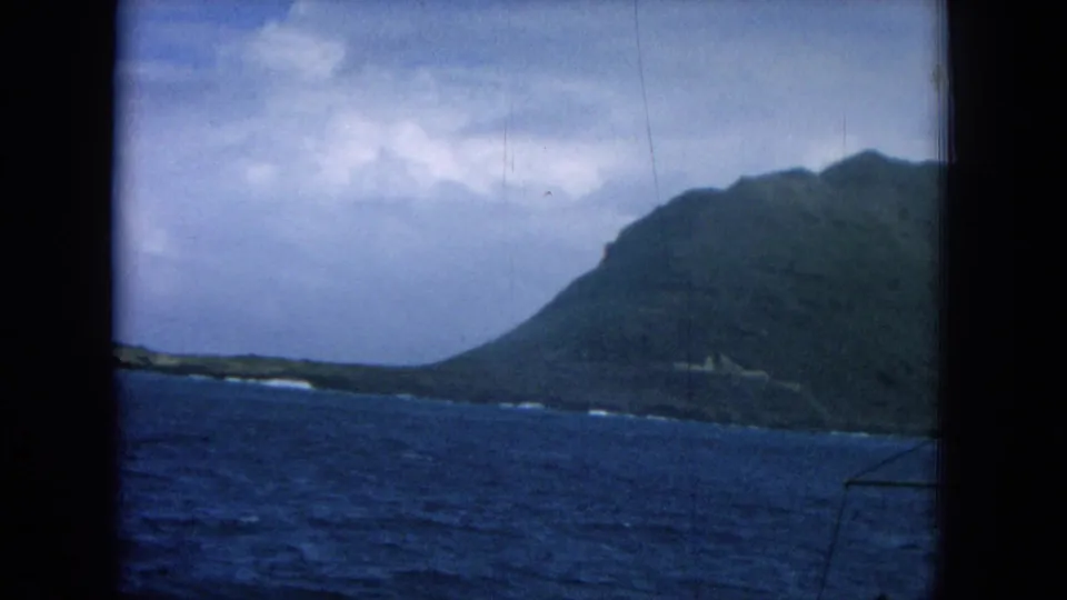 1964 Hawaii home-movie frame showing blue ocean and a steep coastal hill from a moving boat.