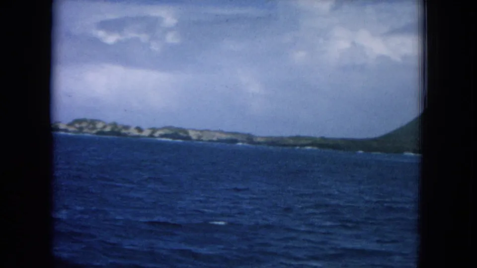 1964 8mm Hawaii travel view of blue water and a distant island coast framed by dark ship window edges.