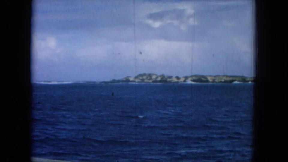 Shipboard 8mm view of a low Hawaiian island across blue ocean, with dark window framing at both sides.