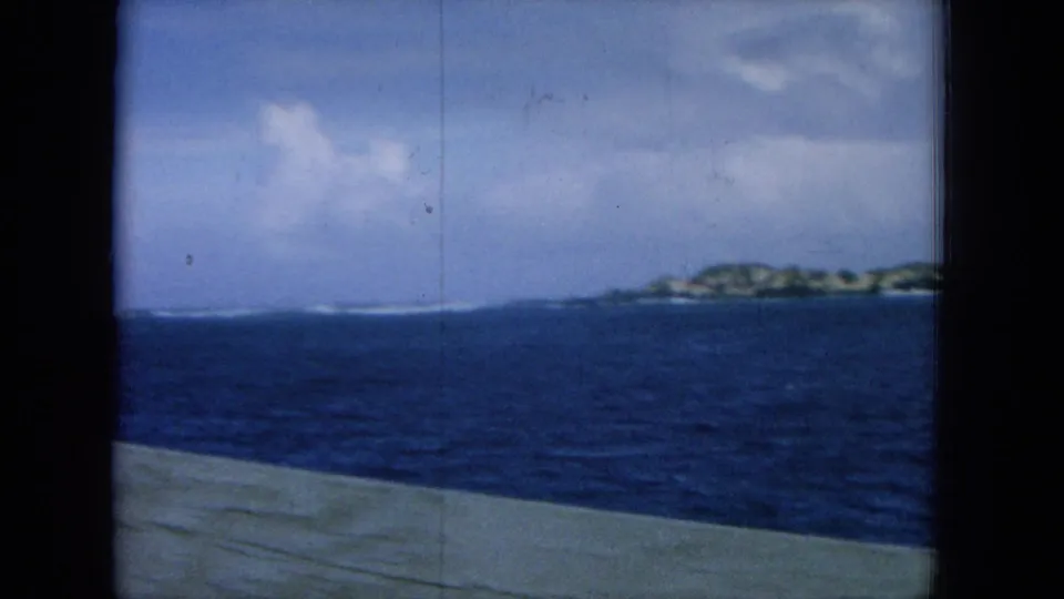 1964 8mm shipboard view showing blue water, island coast, cloudy sky, and a pale deck edge in the foreground.