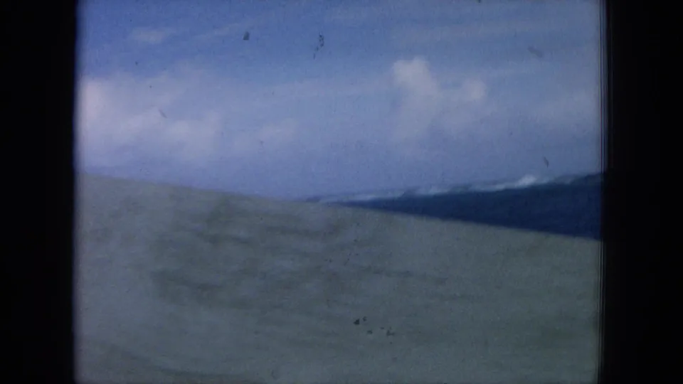 Final shipboard frame with pale deck surface, blue ocean, and a hazy Hawaiian coastline under a cloudy sky.