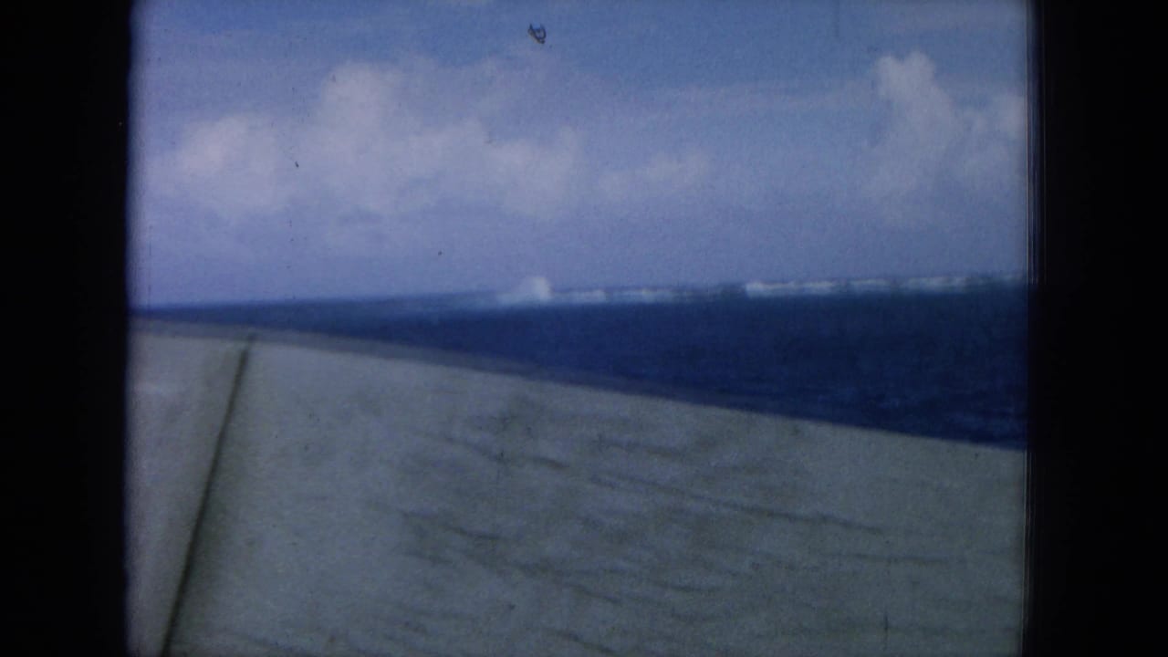 Blue ocean and distant shoreline seen past a ship deck edge and dark vertical frame.