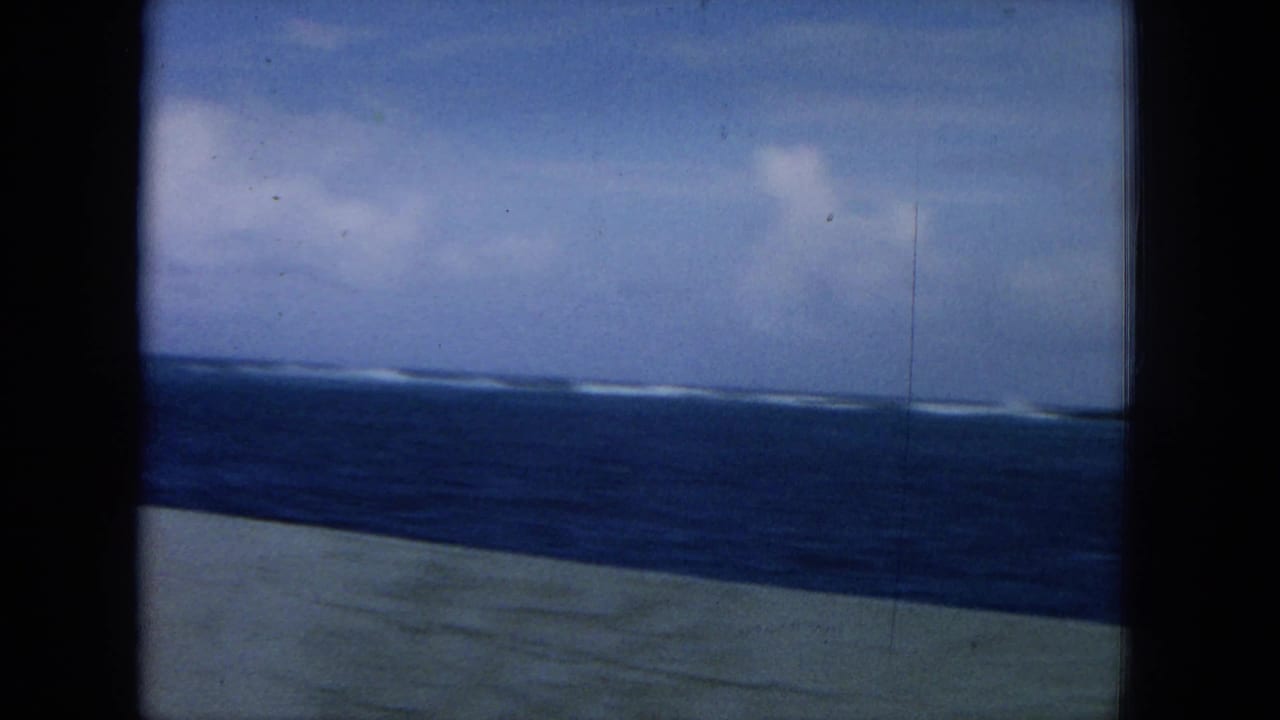 Distant shoreline and surf across blue water, framed by dark vertical bars and a deck edge.