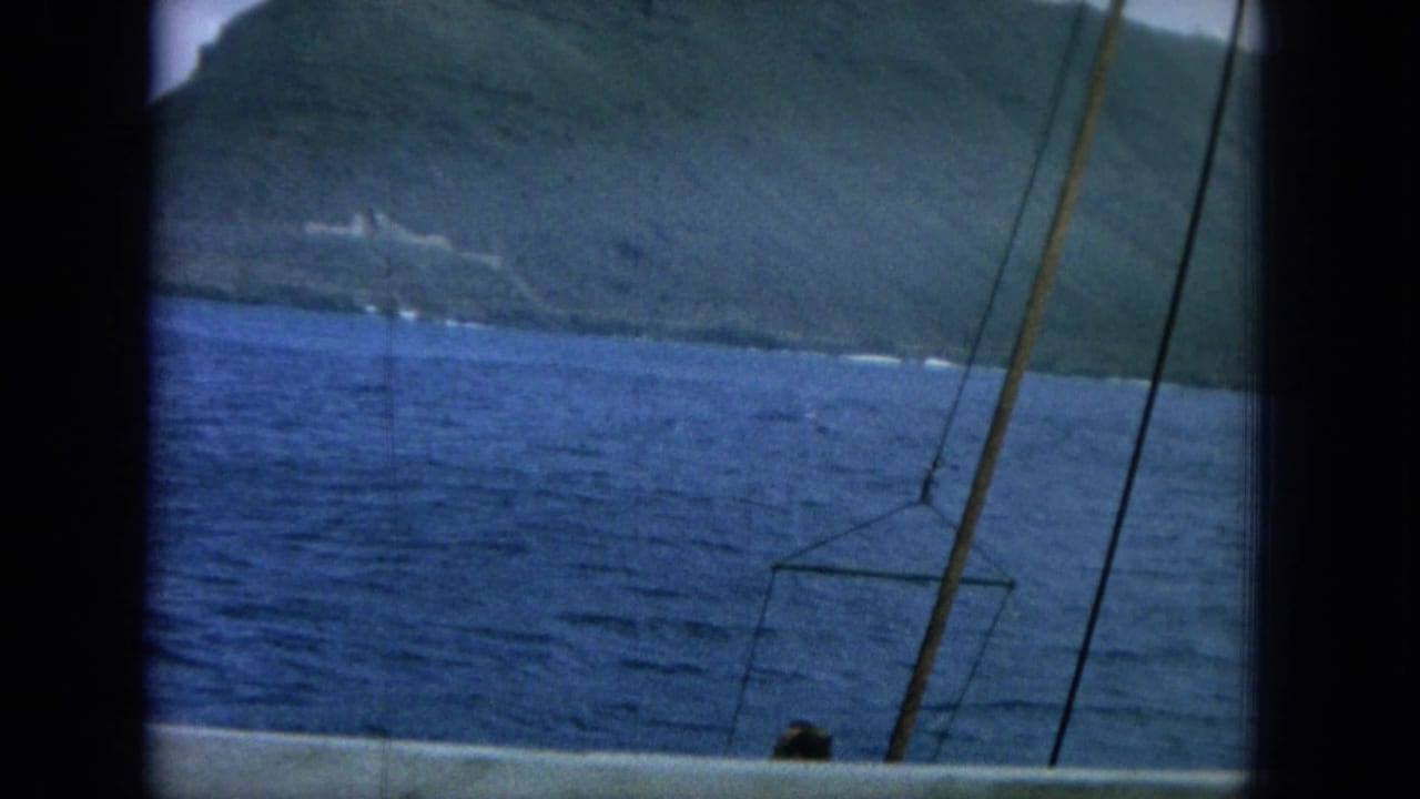 Blue ocean and green mountain shoreline seen from a boat with rigging in the foreground.