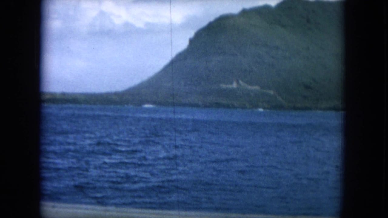 Open water in front of a steep green island slope under a cloudy sky.