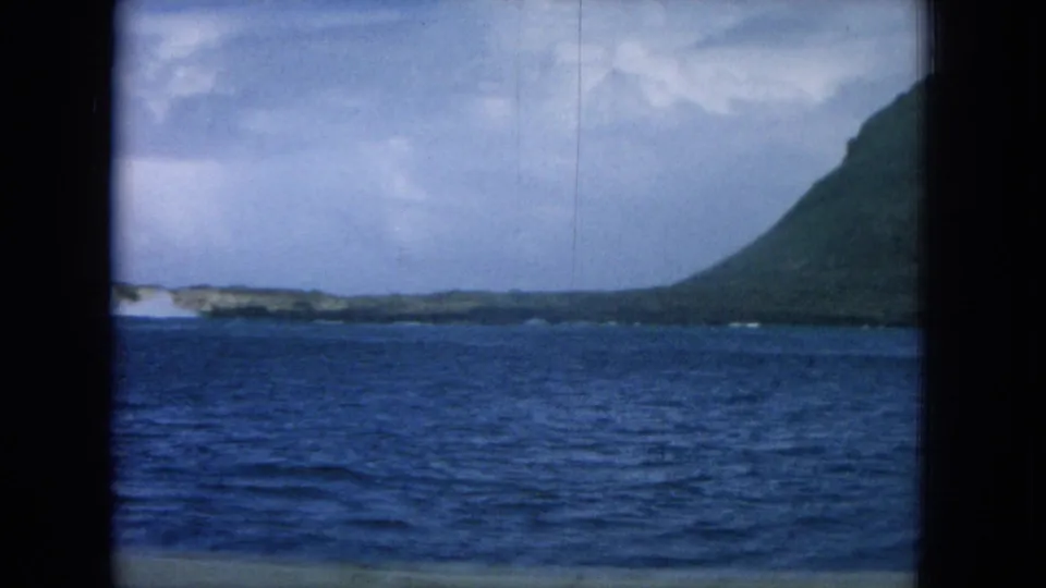 1964 8mm Hawaii boat-view frame showing blue water, low shoreline, surf, and a mountain edge under cloud.