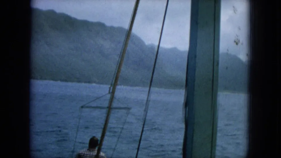1964 8mm Hawaii footage shows a man in a plaid shirt on deck, framed by rigging, looking toward ocean water and mountains.
