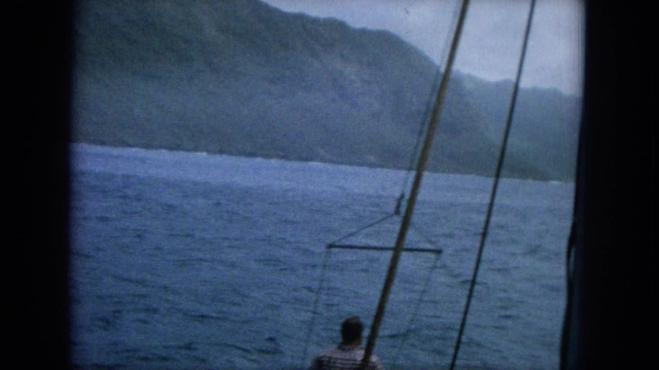Man near boat rigging looking across blue water toward a mountainous coast.